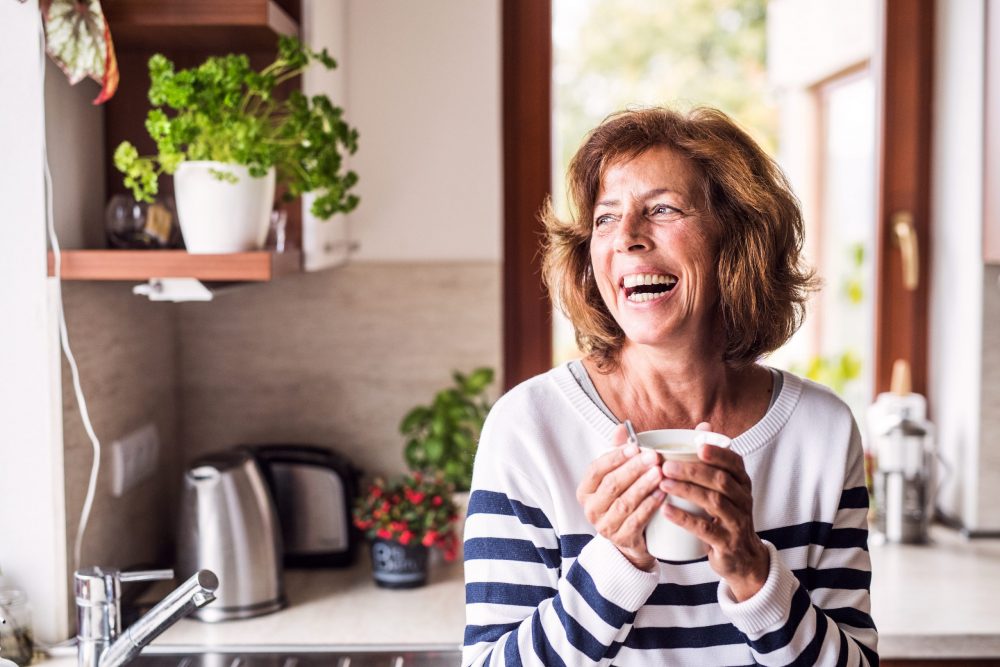 A high-net-worth retired wealth management client holds a coffee cup while laughing in her kitchen.